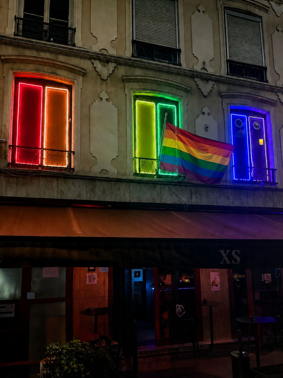 Façade du XS BAR, bar gay à Lyon, avec ses trois fenêtres en néon rouge, vert et bleu et un drapeau arc-en-ciel